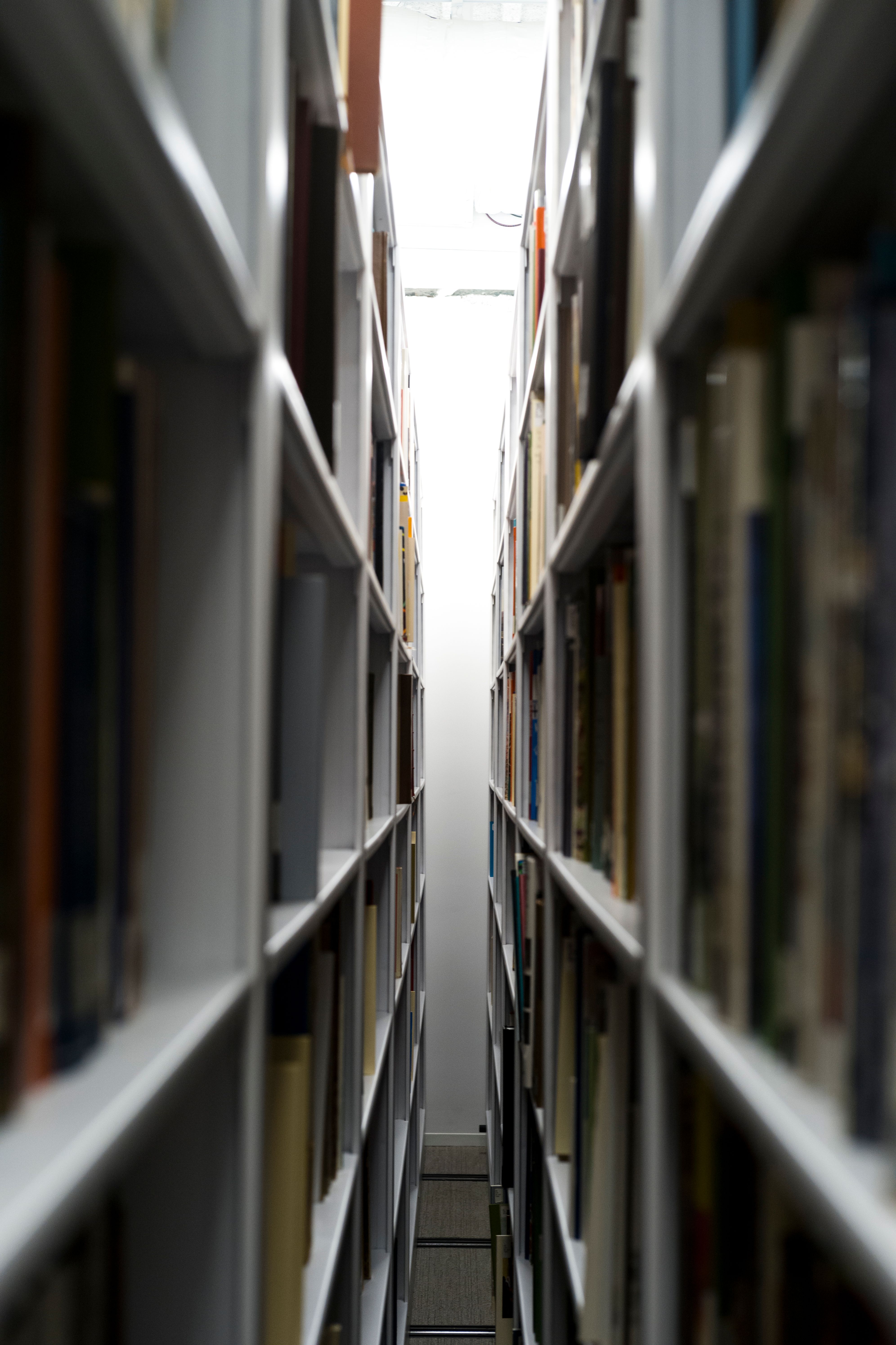 A narrow aisle between tall, fully stocked bookshelves leads to a bright, softly lit space at the end.