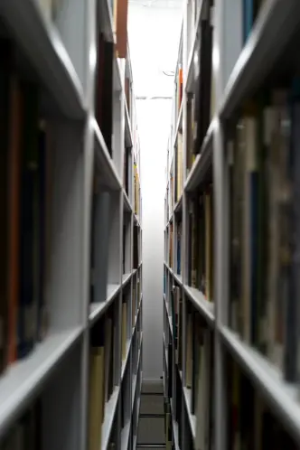 A narrow aisle between tall, fully stocked bookshelves leads to a bright, softly lit space at the end.