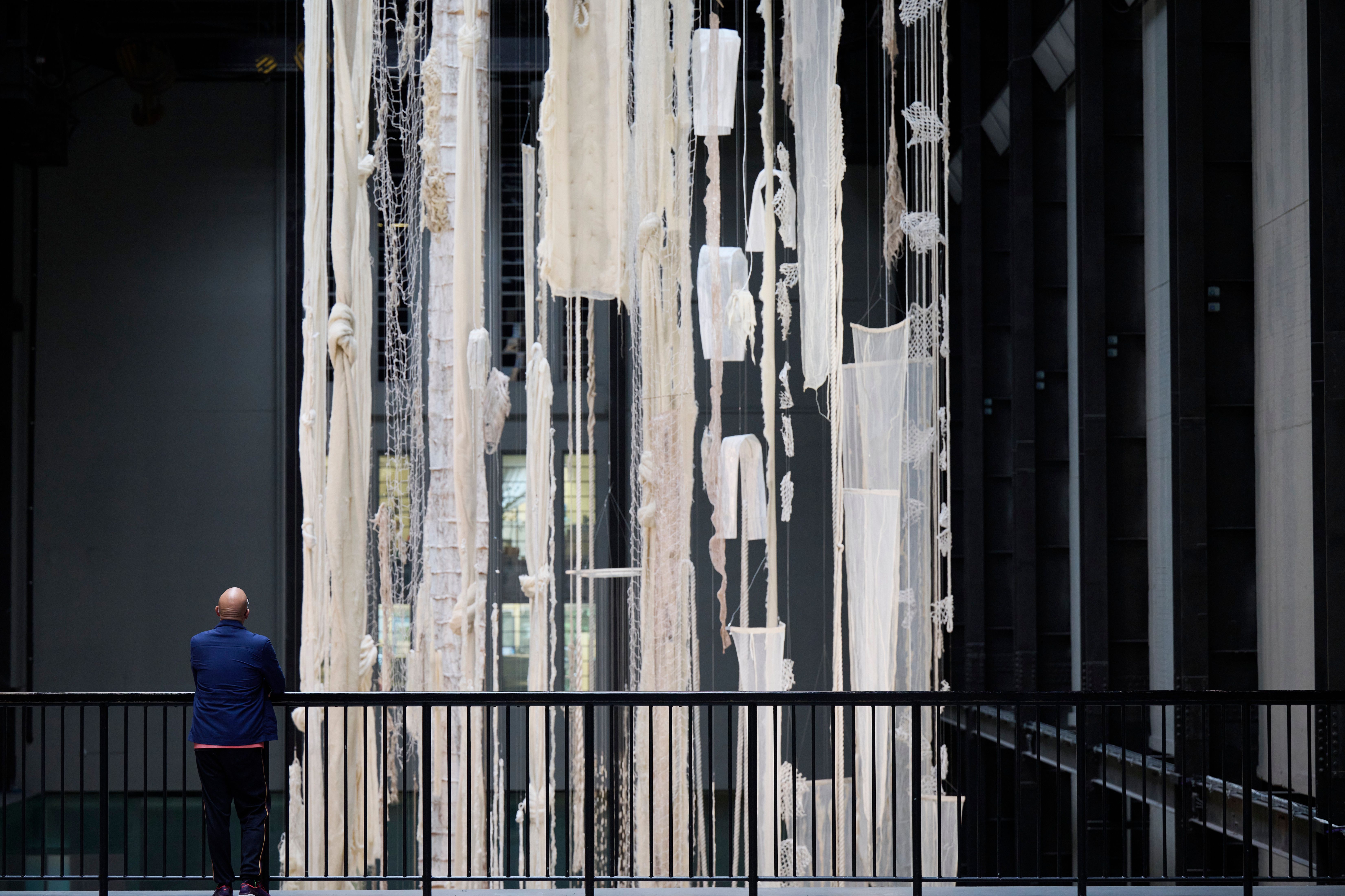 A person leans against a fence, observing suspended white torn fabrics and mesh fishing nets.