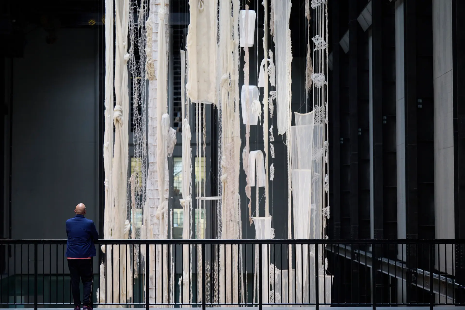 A person leans against a fence, observing suspended white torn fabrics and mesh fishing nets.