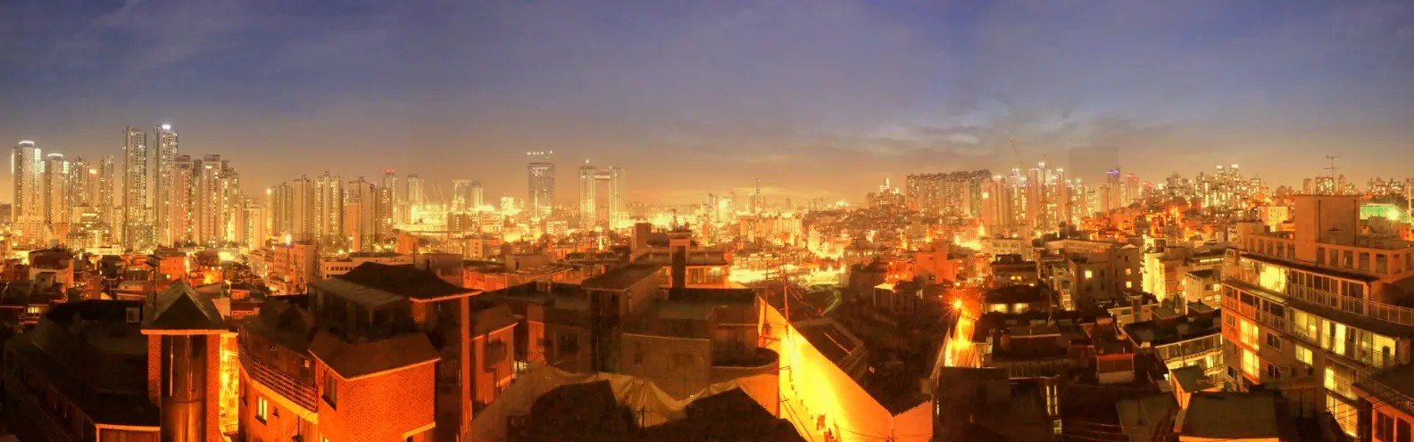 A panoramic view of a city at night, illuminated by a warm, orange glow against a deep blue sky, captured in a still from ...