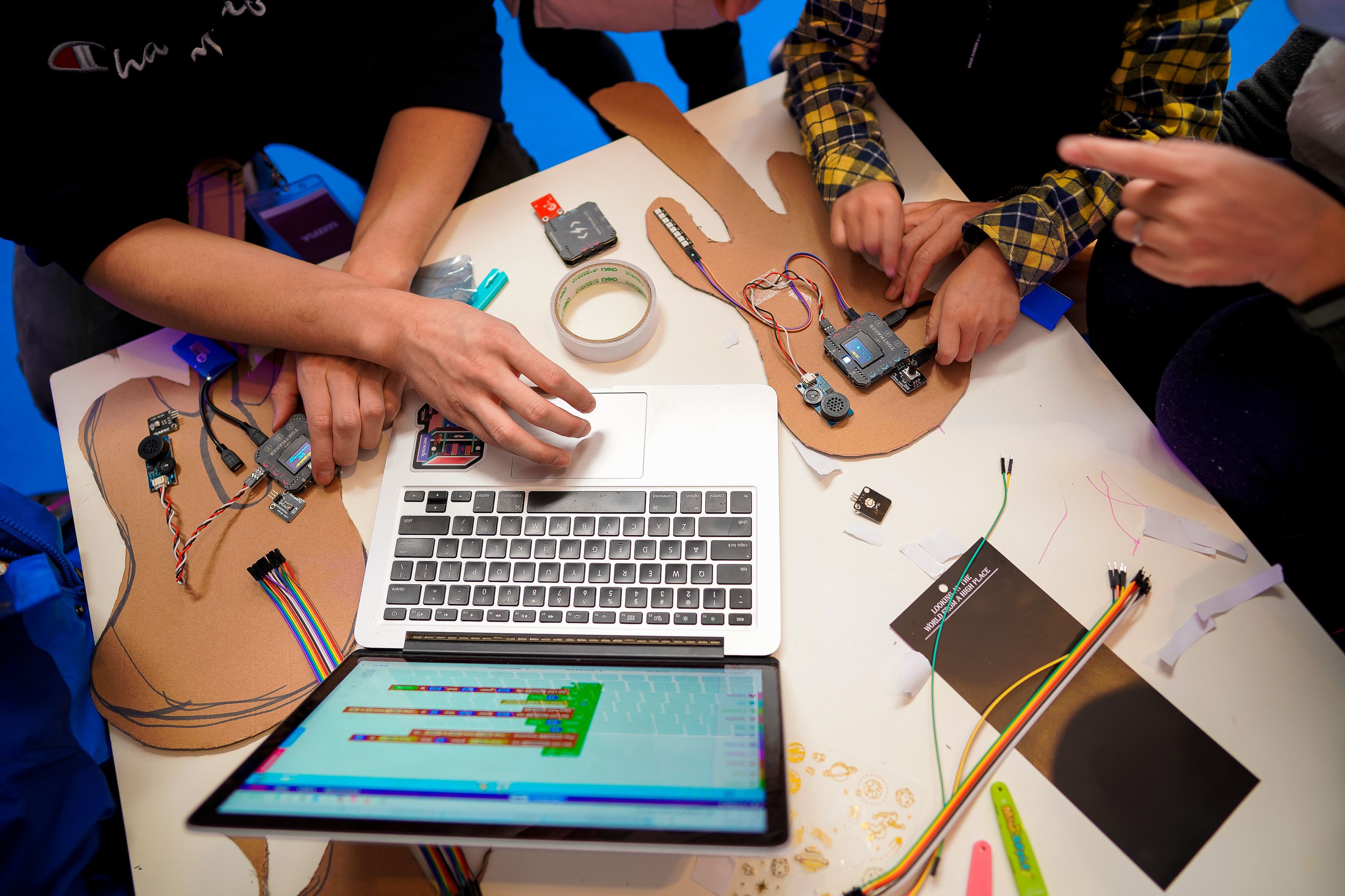 An overhead view of a desk featuring guitar prototypes, a laptop, and two pairs of hands interacting with the prototypes a...