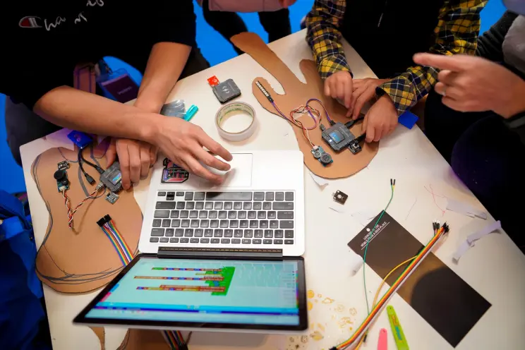 An overhead view of a desk featuring guitar prototypes, a laptop, and two pairs of hands interacting with the prototypes a...