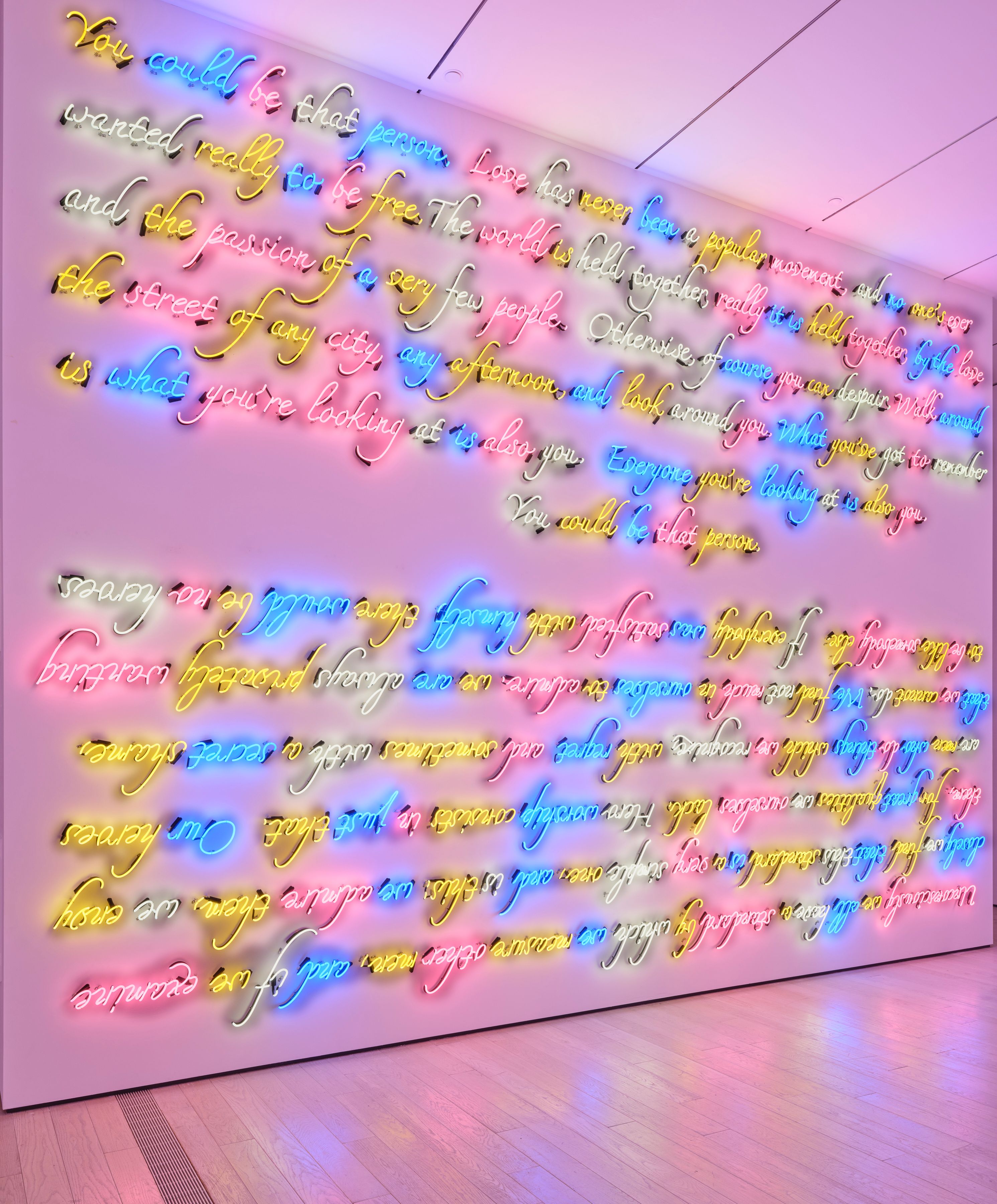 Bright multicolored neon cursive text covering an entire gallery wall, glowing pink, yellow, blue, and white.