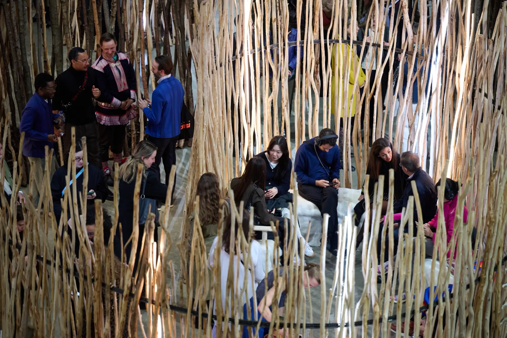 Visitors sit and converse inside a circular wooden installation, surrounded by vertical branches and integrated lighting.
