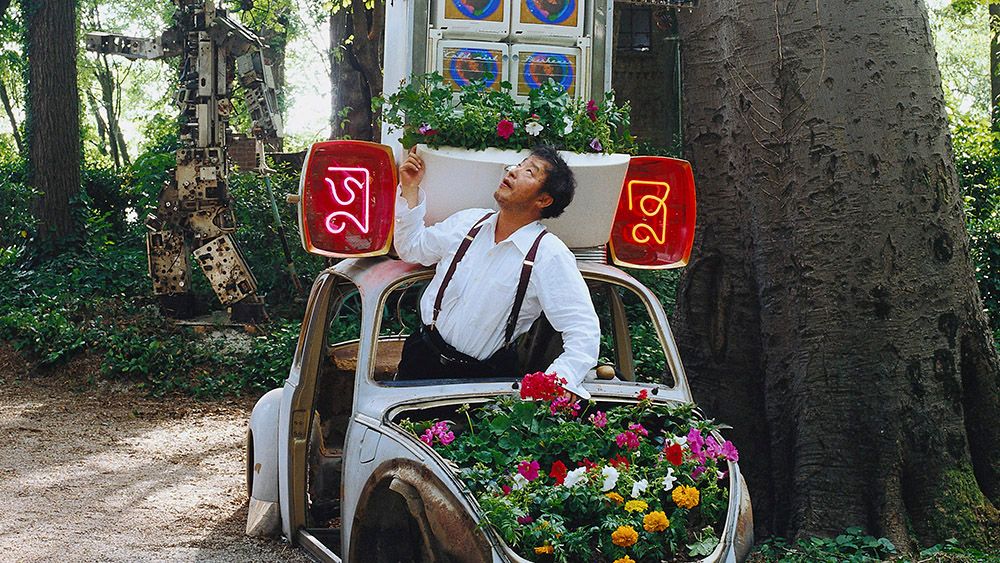 A man stands in a flower-filled vintage car with neon signs, looking up. The scene is set in a lush wooded area with sculp...