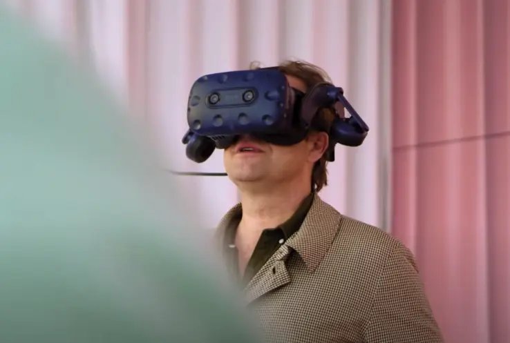 A person donned in a virtual reality headset stands against a backdrop of pink walls, their expression showing amusement.