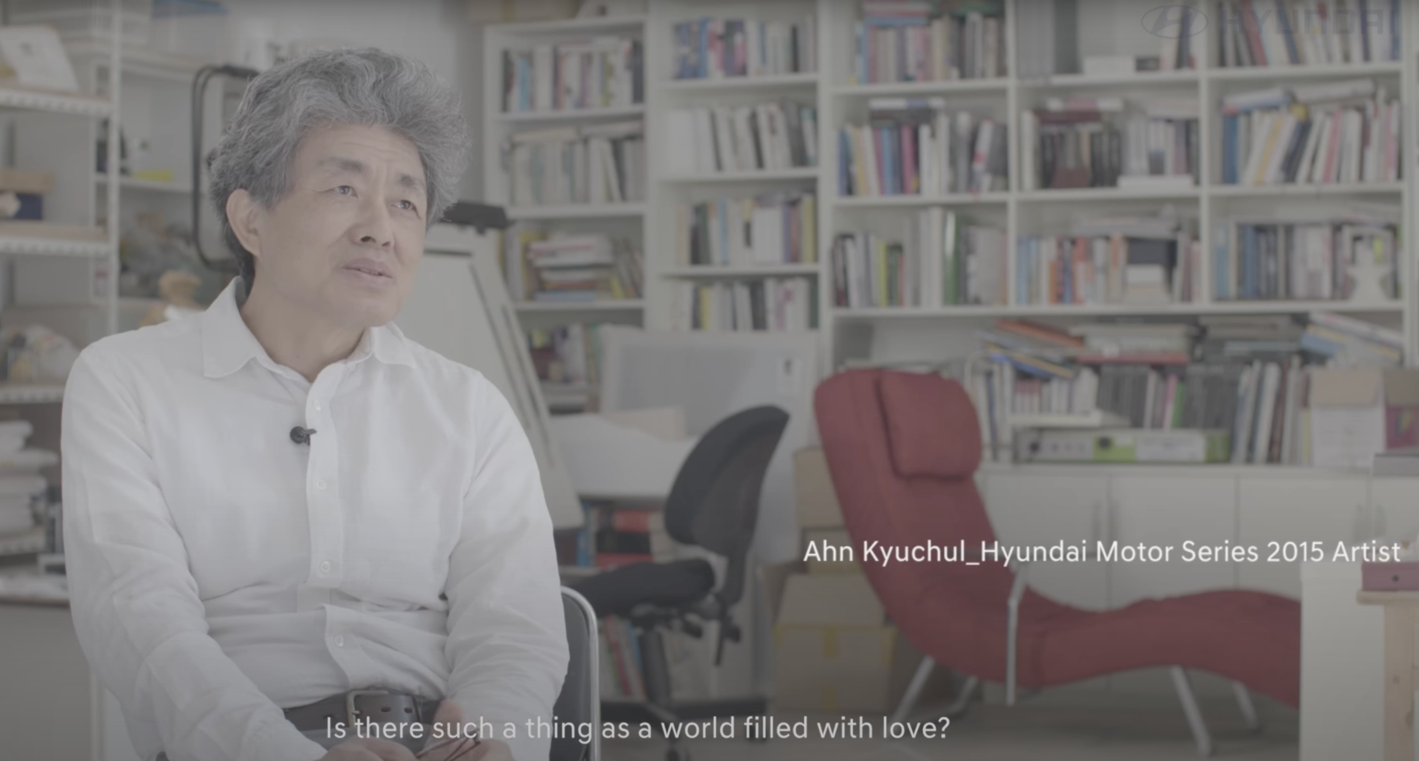 A person sitting in an office with a microphone clipped to their white shirt, in front of bookshelves filled with books. B...