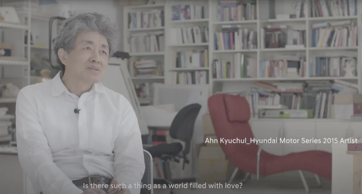A person sitting in an office with a microphone clipped to their white shirt, in front of bookshelves filled with books. Behind them are two office chairs. The text "Ahn Kyuchul_Hyundai Motor Series 2015 Artist" is displayed prominently.