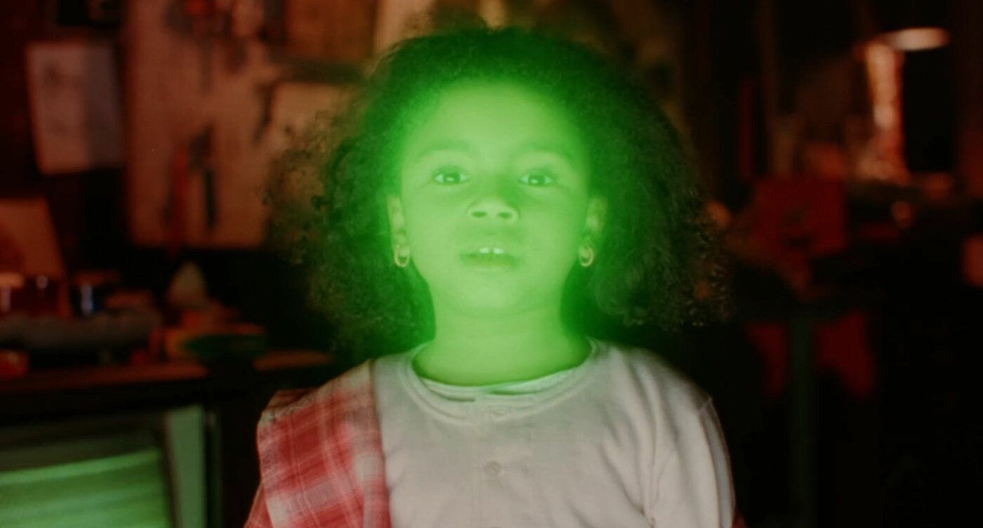 A young girl with a glowing green aura around her face stands in a dimly lit room.