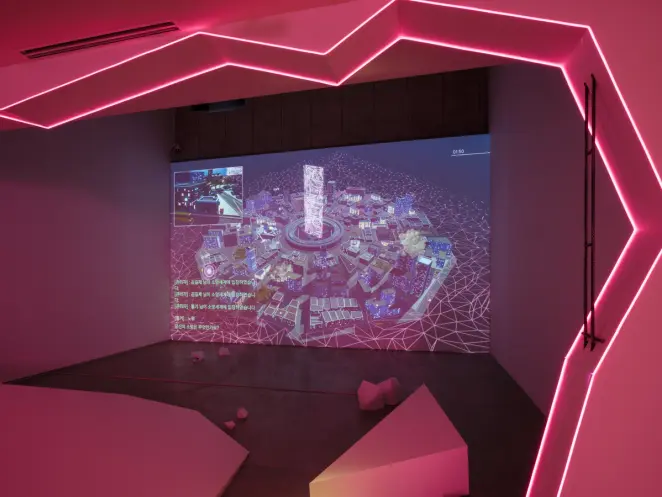A darkened gallery lit by pink neon outlining a jagged hexagonal frame. A digital projection of a virtual cityscape with K...