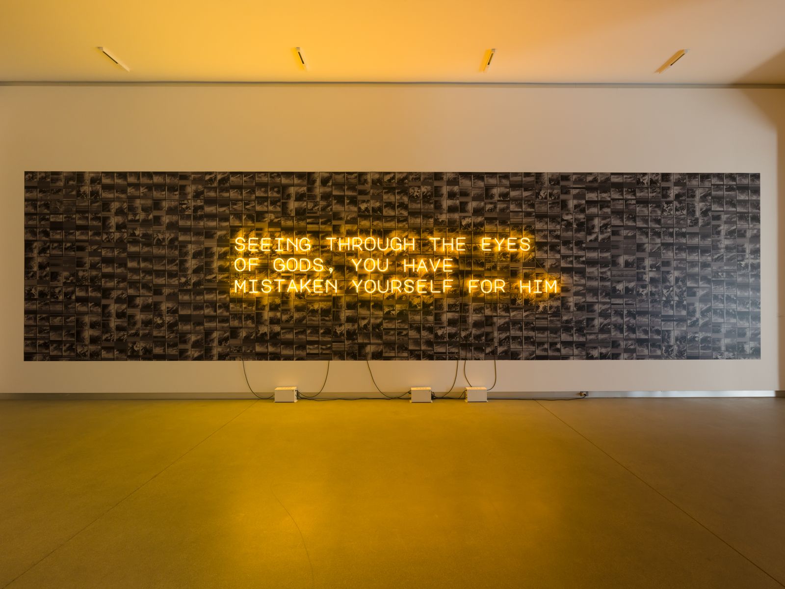 Large wall installation with yellow neon text reading “SEEING THROUGH THE EYES OF GODS, YOU HAVE MISTAKEN YOURSELF FOR HIM” over a grid of black-and-white images.