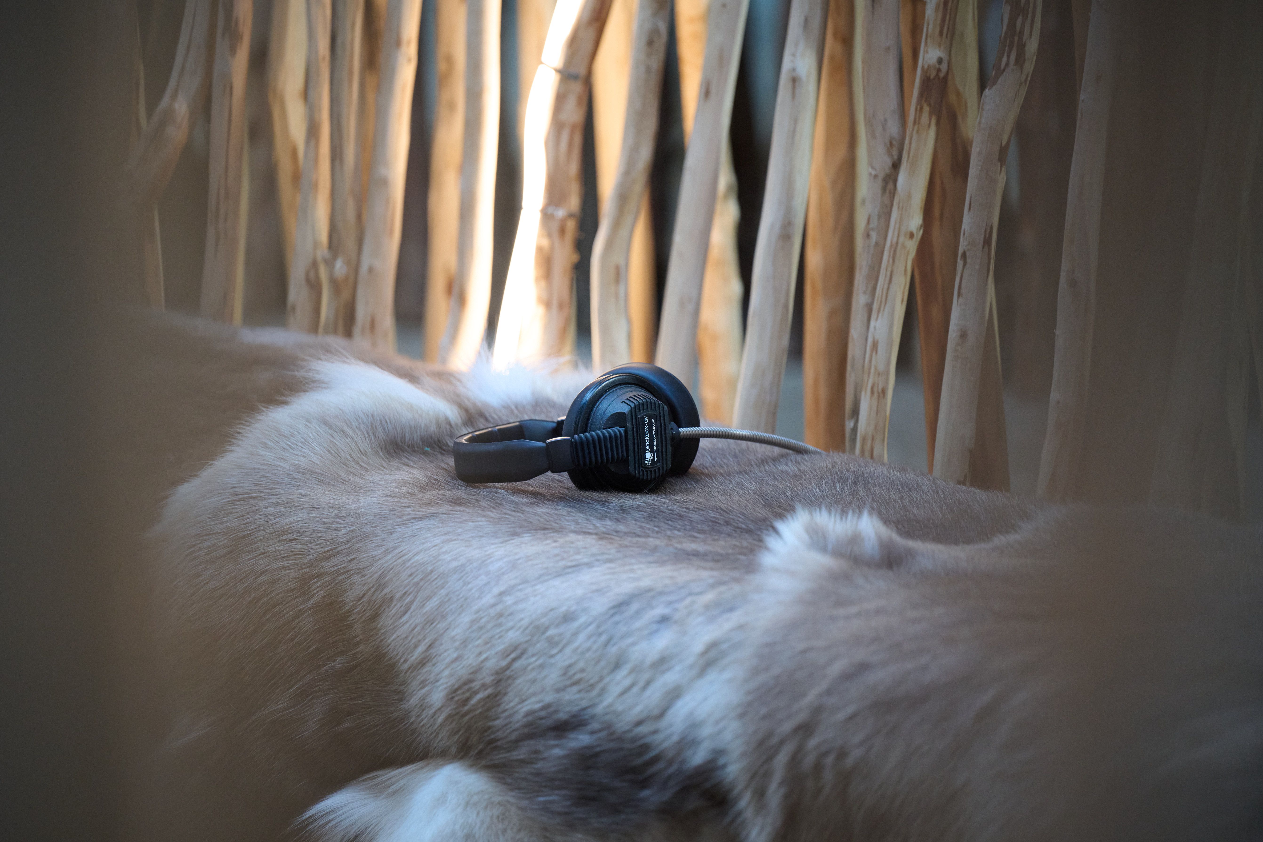 A pair of black over-ear headphones rests on a pale animal hide laid over a wooden structure in soft natural light.