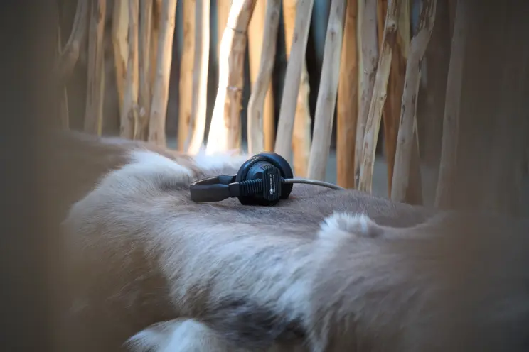A pair of black over-ear headphones rests on a pale animal hide laid over a wooden structure in soft natural light.