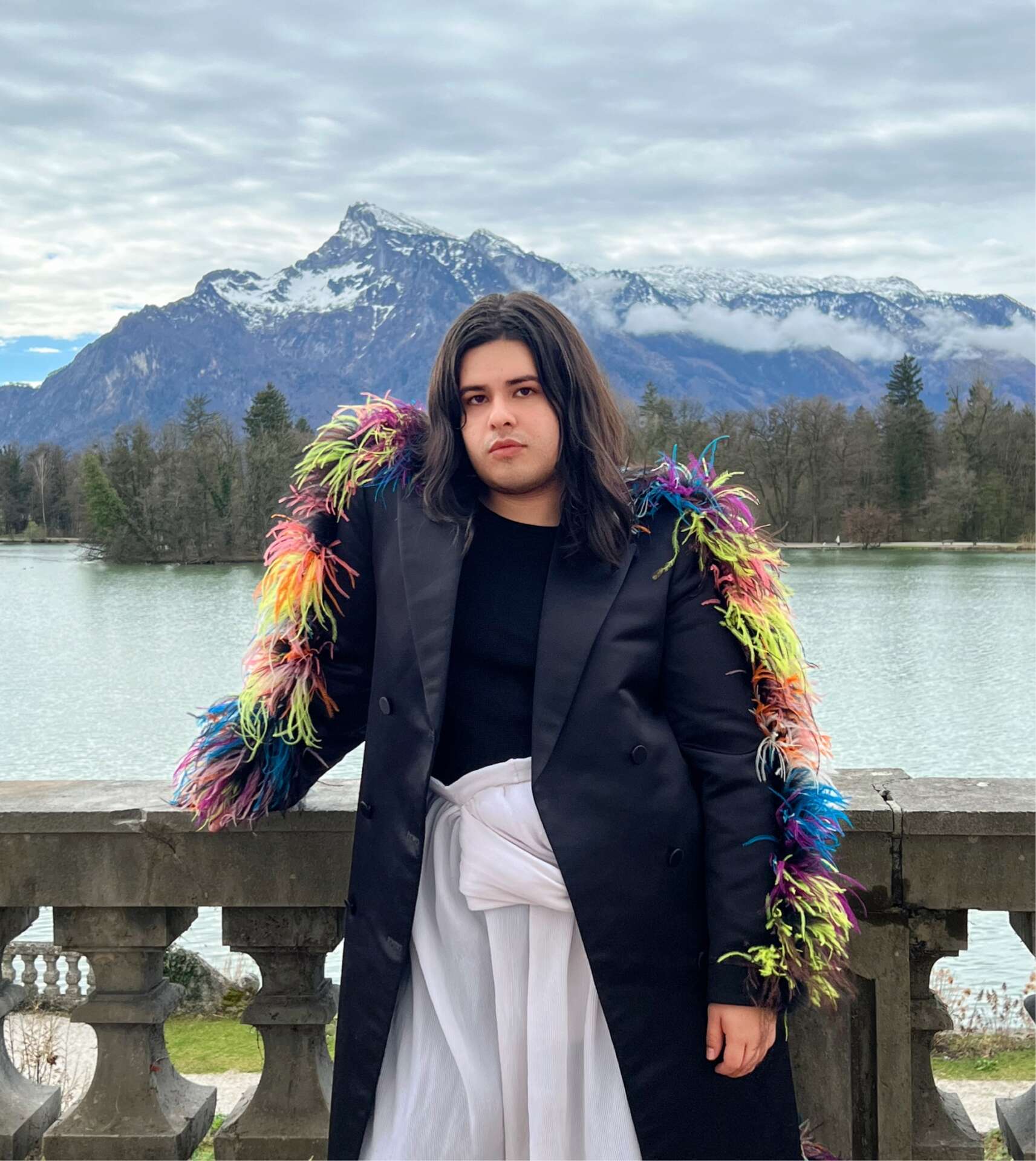Portrait of a person with dark hair, wearing a black coat adorned with colorful feather-like decorations, standing in fron...