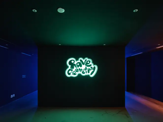 A black canvas in a dark room illuminated by backlights in hues of blue, green, and red, showcasing geometric shapes and u...