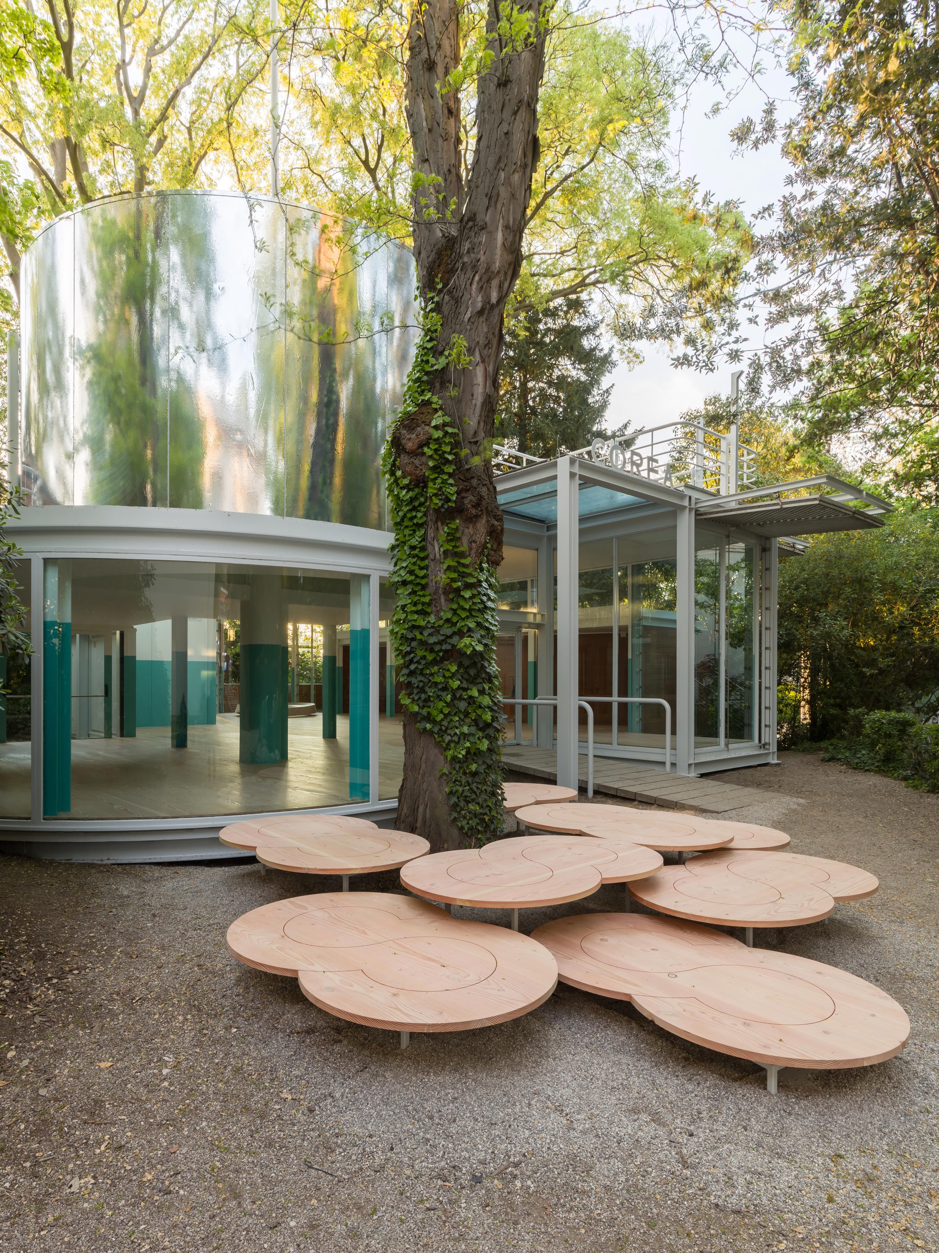 A view of a contemporary pavilion surrounded by trees, with a mirrored section and wooden tables.