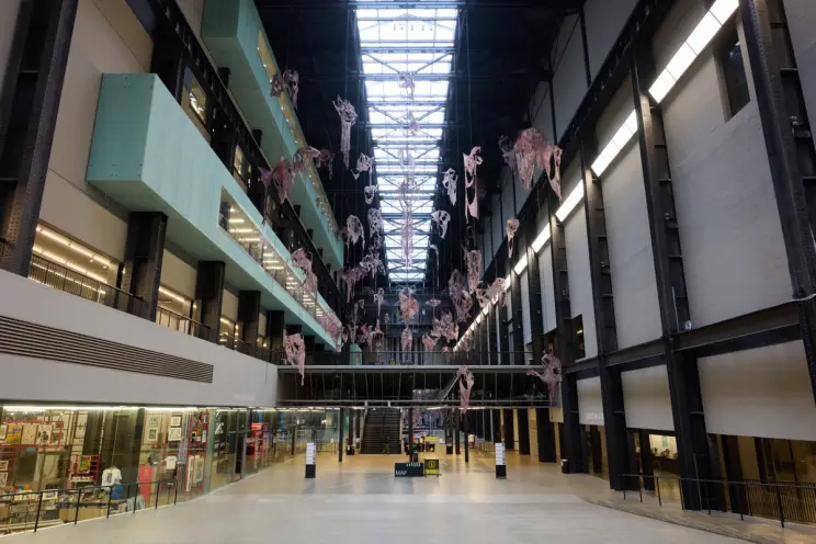 Wide view of industrial gallery space with hanging fleshy sculptures, large skylight overhead.