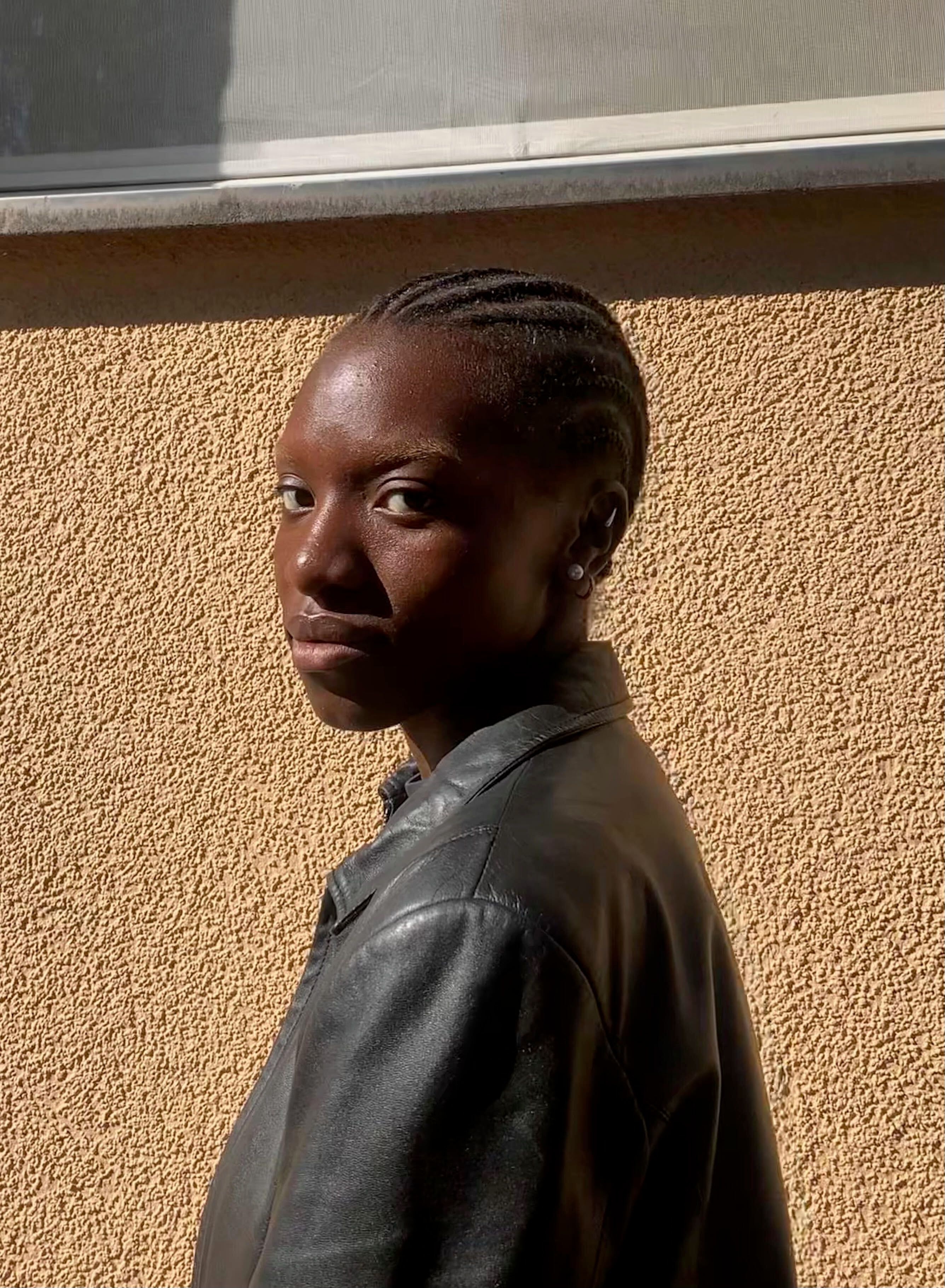 A person with short, braided hair stands against a textured wall, wearing a black leather jacket and looking directly at t...