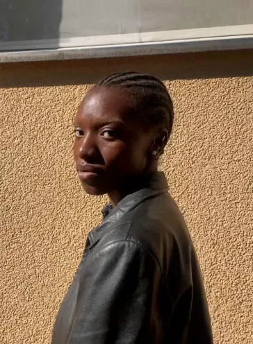 A person with short, braided hair stands against a textured wall, wearing a black leather jacket and looking directly at t...
