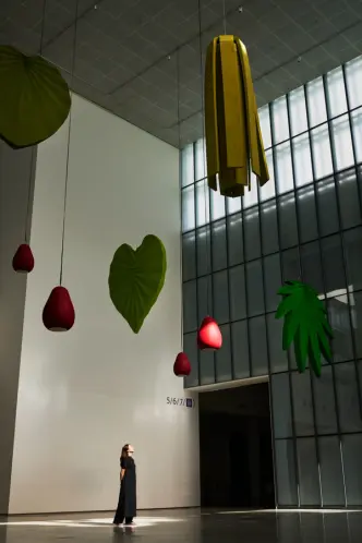 Large hanging leaf and fruit sculptures in a sunlit atrium with a person looking up at the artwork.