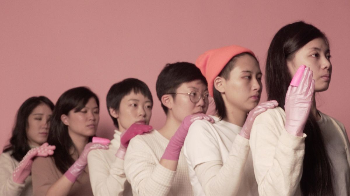 Six individuals in a line on a vivid pink background, each wearing pink gloves and resting their hands on the shoulder of the person in front. The person at the front extends their palm outward.