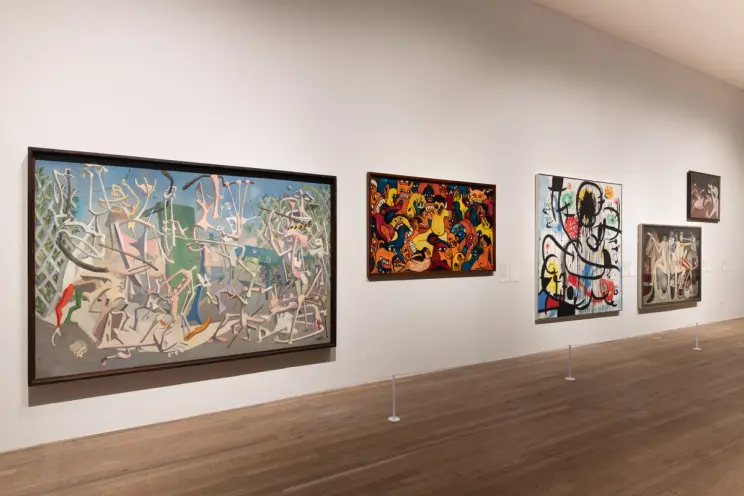 A variety of abstract art paintings in frames hung on the gallery wall, with a view of the wooden floor beneath.