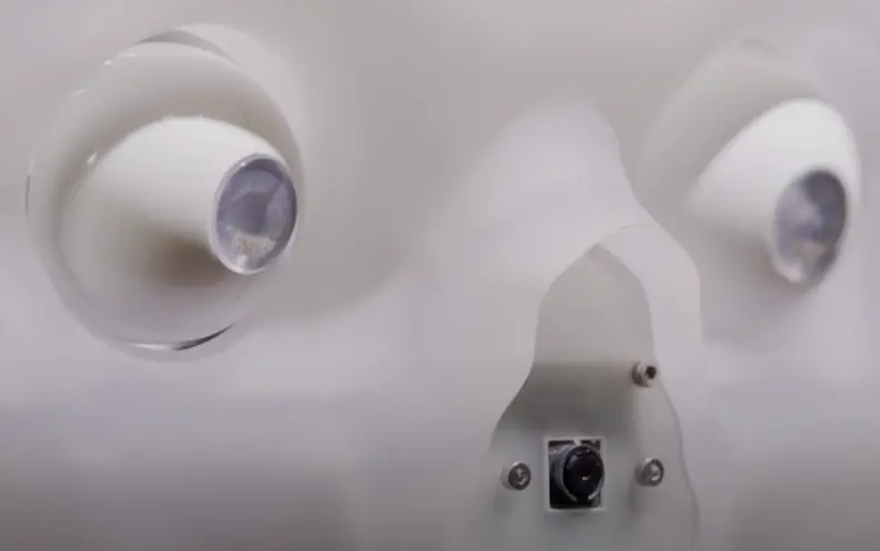 A section of a white mechanical device, featuring two circular lenses positioned side-by-side and a black button in the mi...