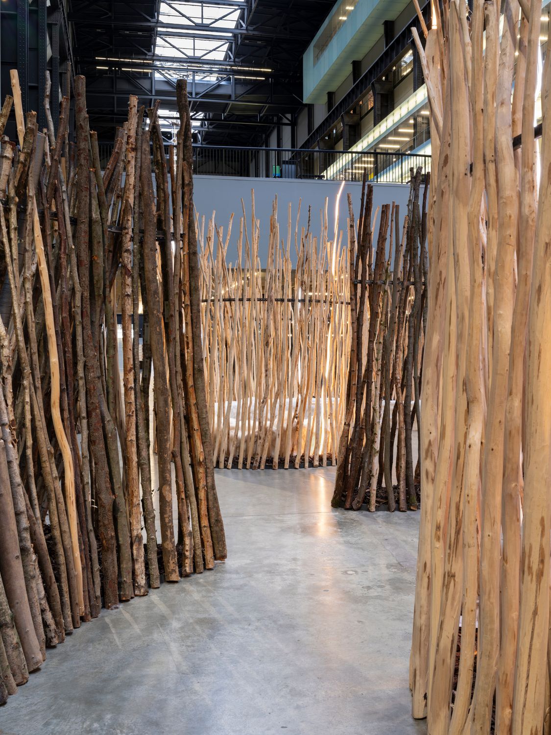 A passageway between tall, rough wooden branch walls leads into a softly illuminated installation inside an industrial hall.