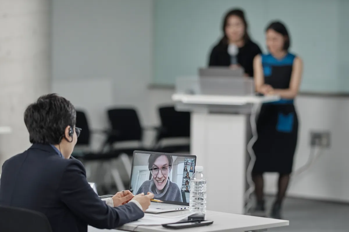 An individual engaging in a video conference at a desk, with two blurred figures standing at a lectern in the background.