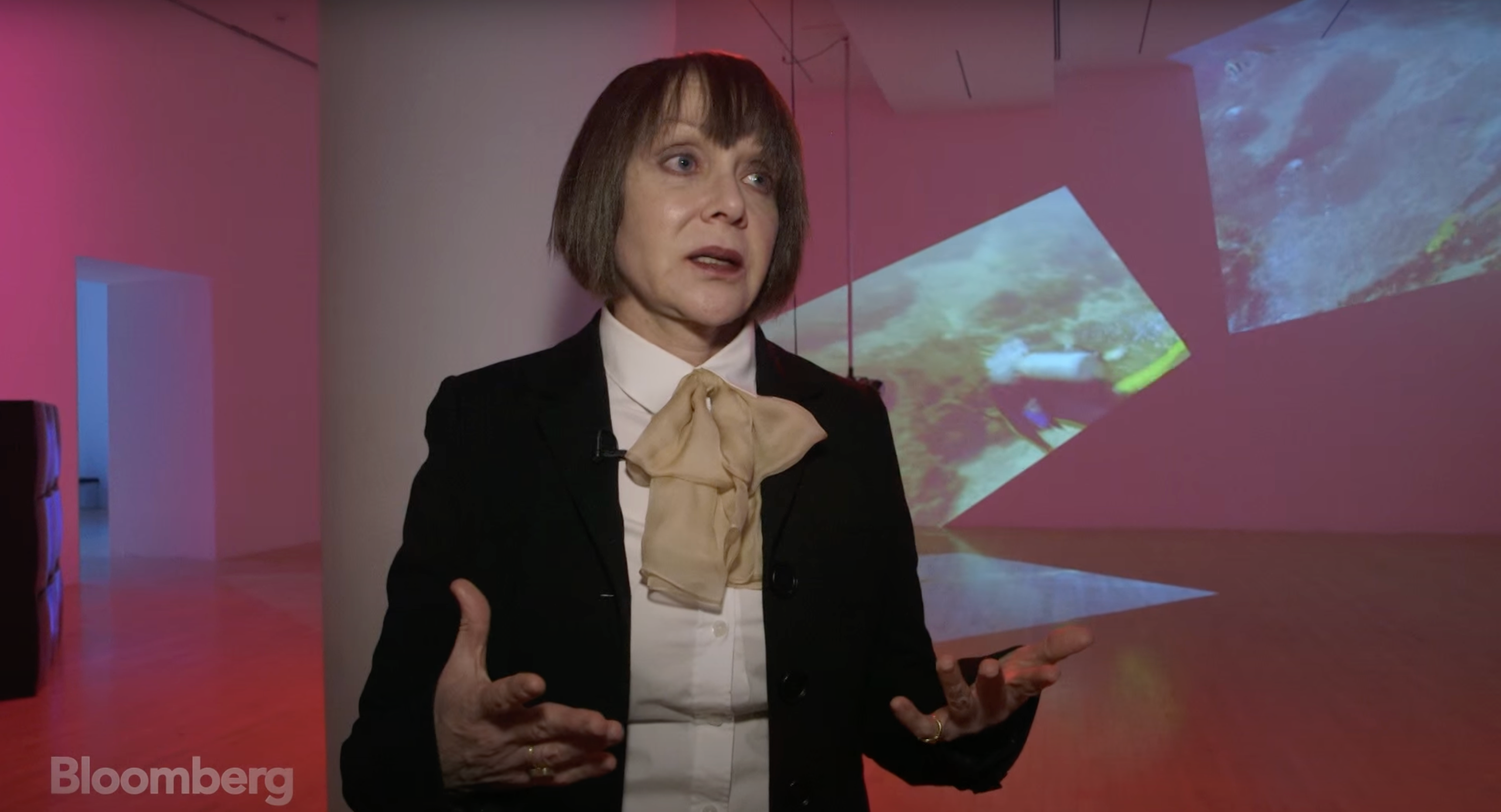 A person is addressing the camera and making a gesture with their hand, standing in front of an illuminated artwork projec...