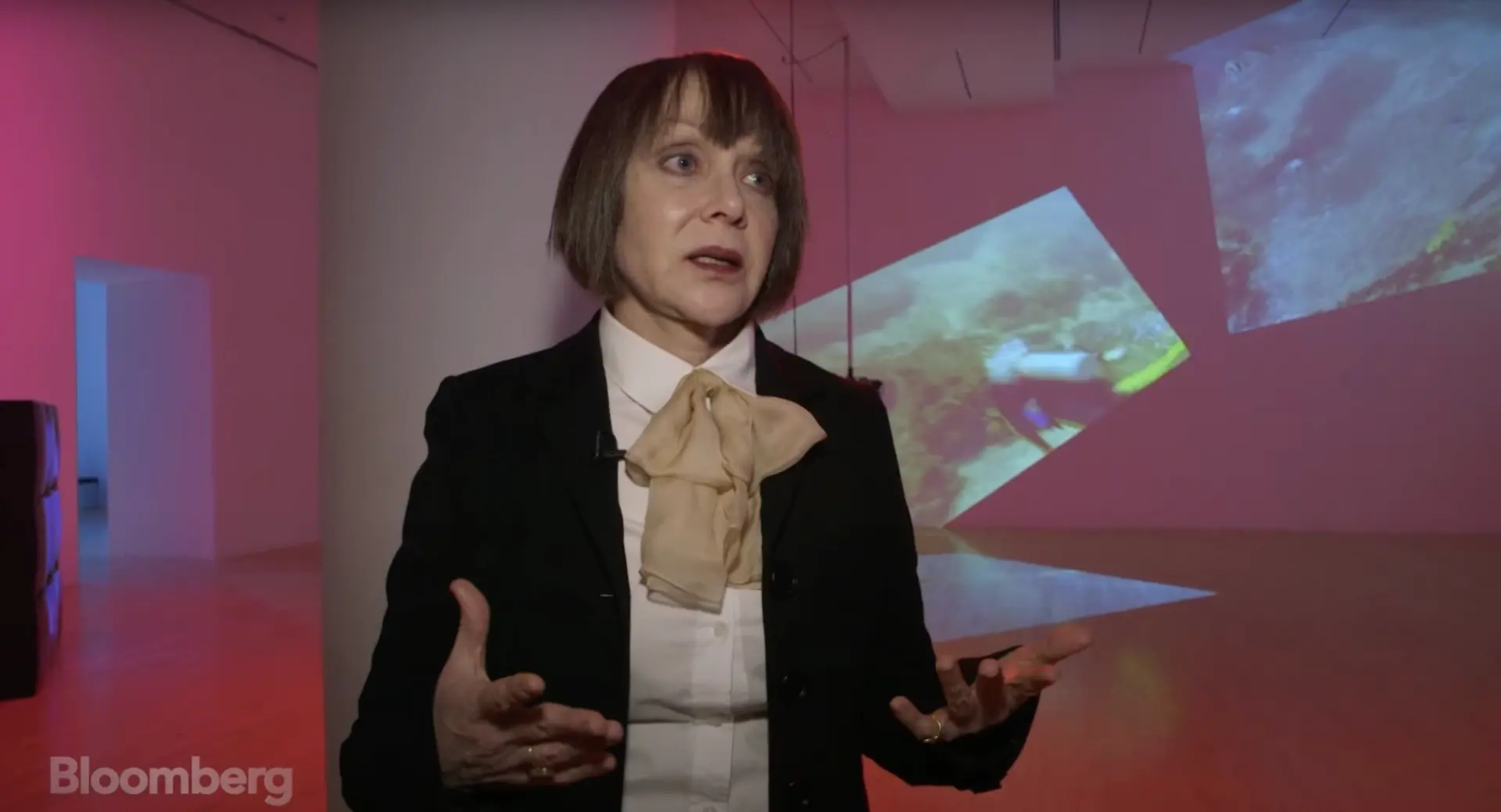 A person is addressing the camera and making a gesture with their hand, standing in front of an illuminated artwork projec...
