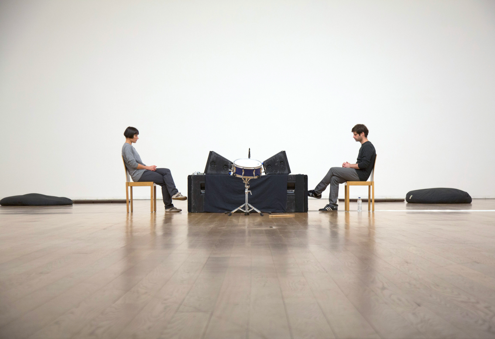 Two individuals sit in wooden chairs on a polished wooden floor, attentively facing each other with drums and black object...
