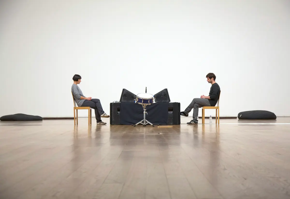 Two individuals sit in wooden chairs on a polished wooden floor, attentively facing each other with drums and black objects positioned between them.