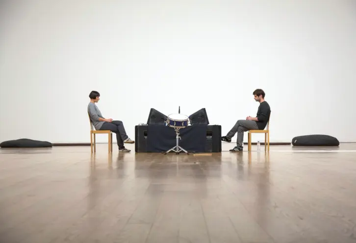 Two individuals sit in wooden chairs on a polished wooden floor, attentively facing each other with drums and black object...