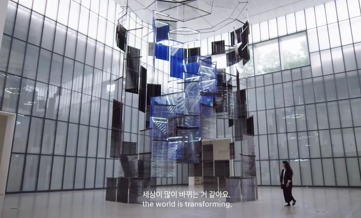 An art installation of rectangular panels in black and blue beginning to form a cylindrical shape, hanging from the ceiling. A person is observing the artwork. The phrase 'The world is transforming' is visible.