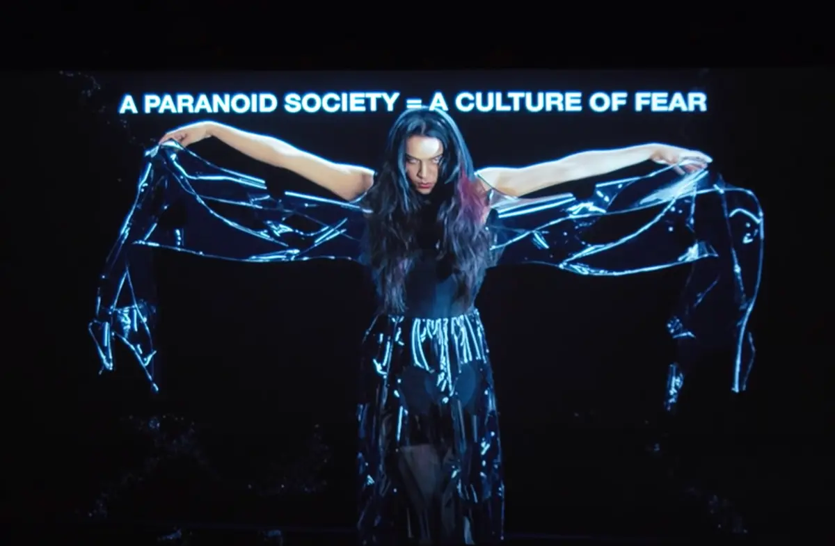 An individual with long black hair wearing an all-black ensemble, spreading out dark fabric attached to their suit reminiscent of spreading wings. The stark white text in the foreground reads, "a paranoid society=a culture of fear."