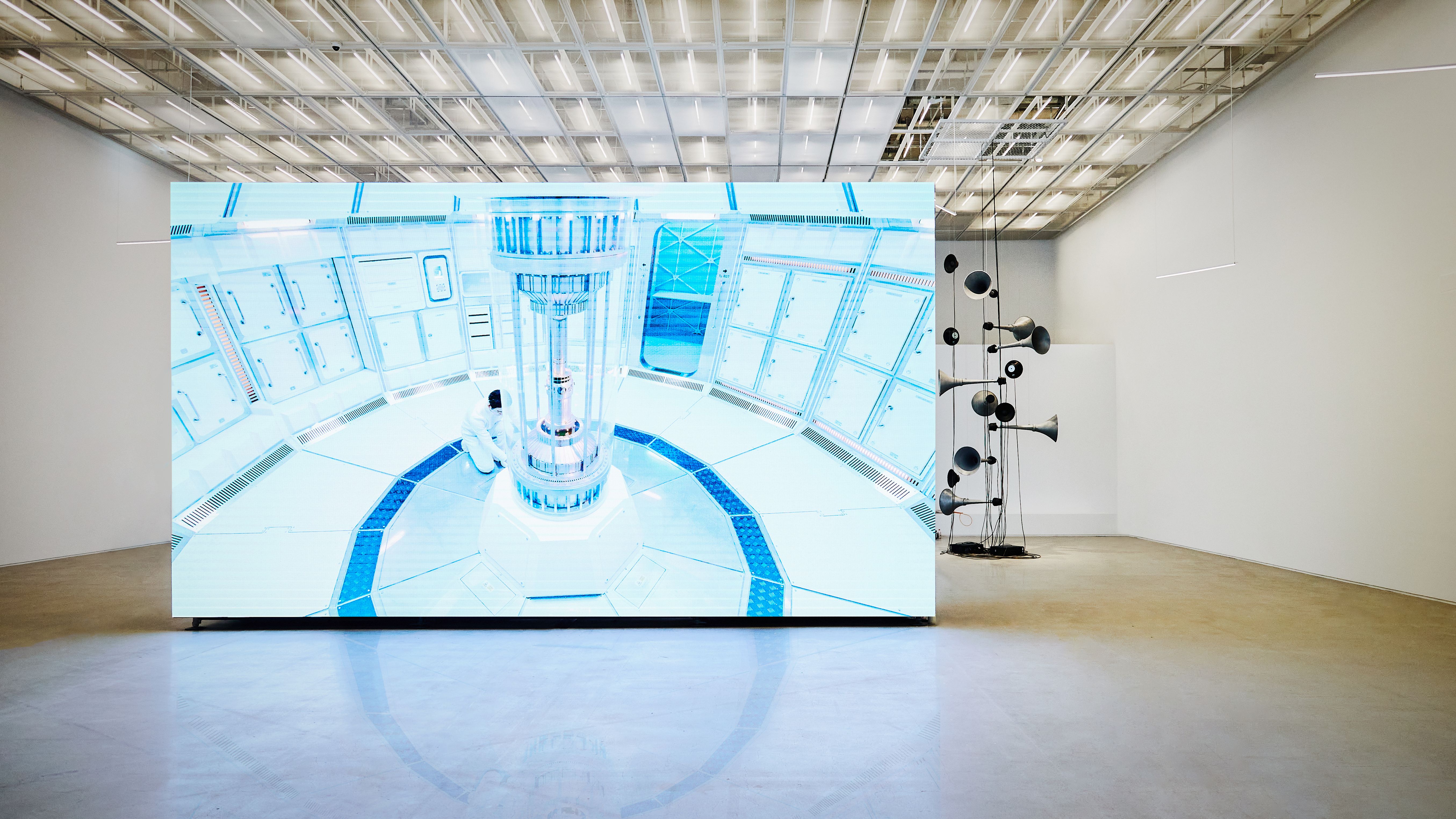 A large gallery space showcasing a two-channel HD film installation. The screen displays a vibrant, sci-fi scene.