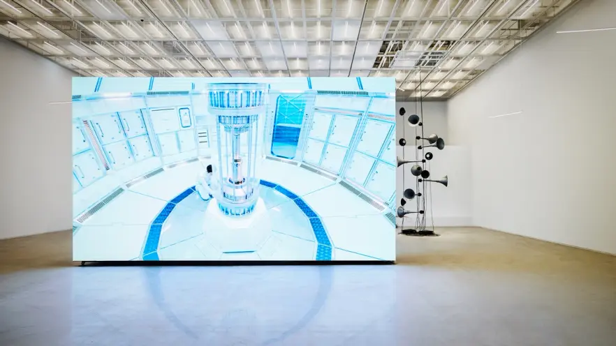 A large gallery space showcasing a two-channel HD film installation. The screen displays a vibrant, sci-fi scene.