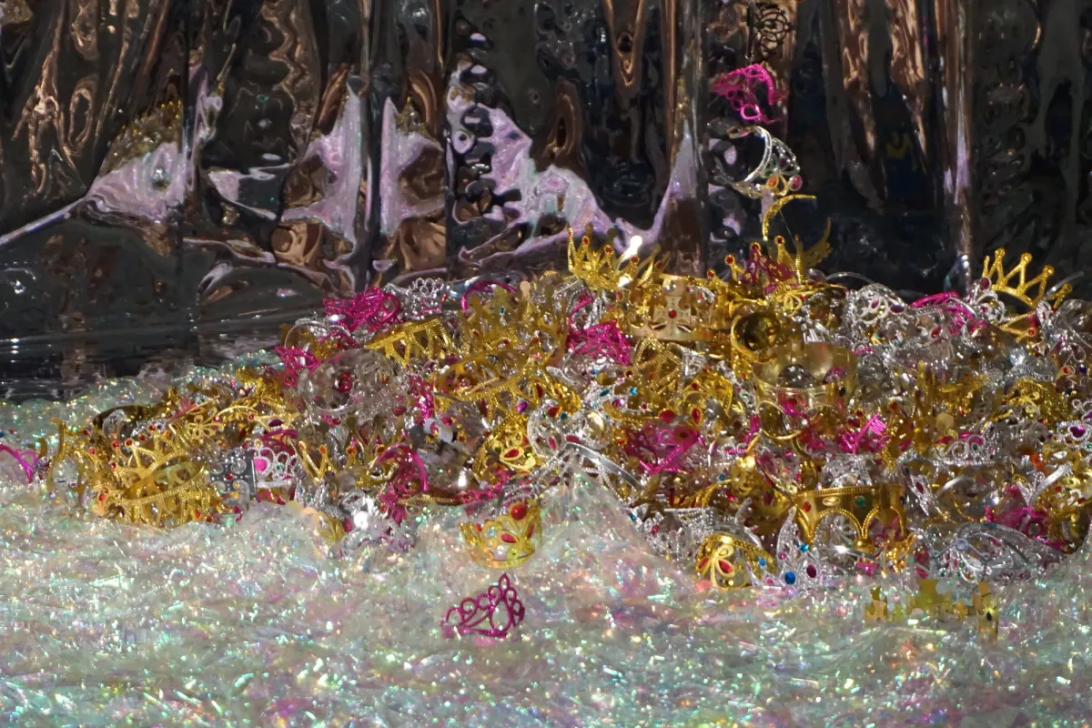 A pile of golden, silver, and pink crowns atop a white shimmering material, against a dark, shiny, uneven structure.