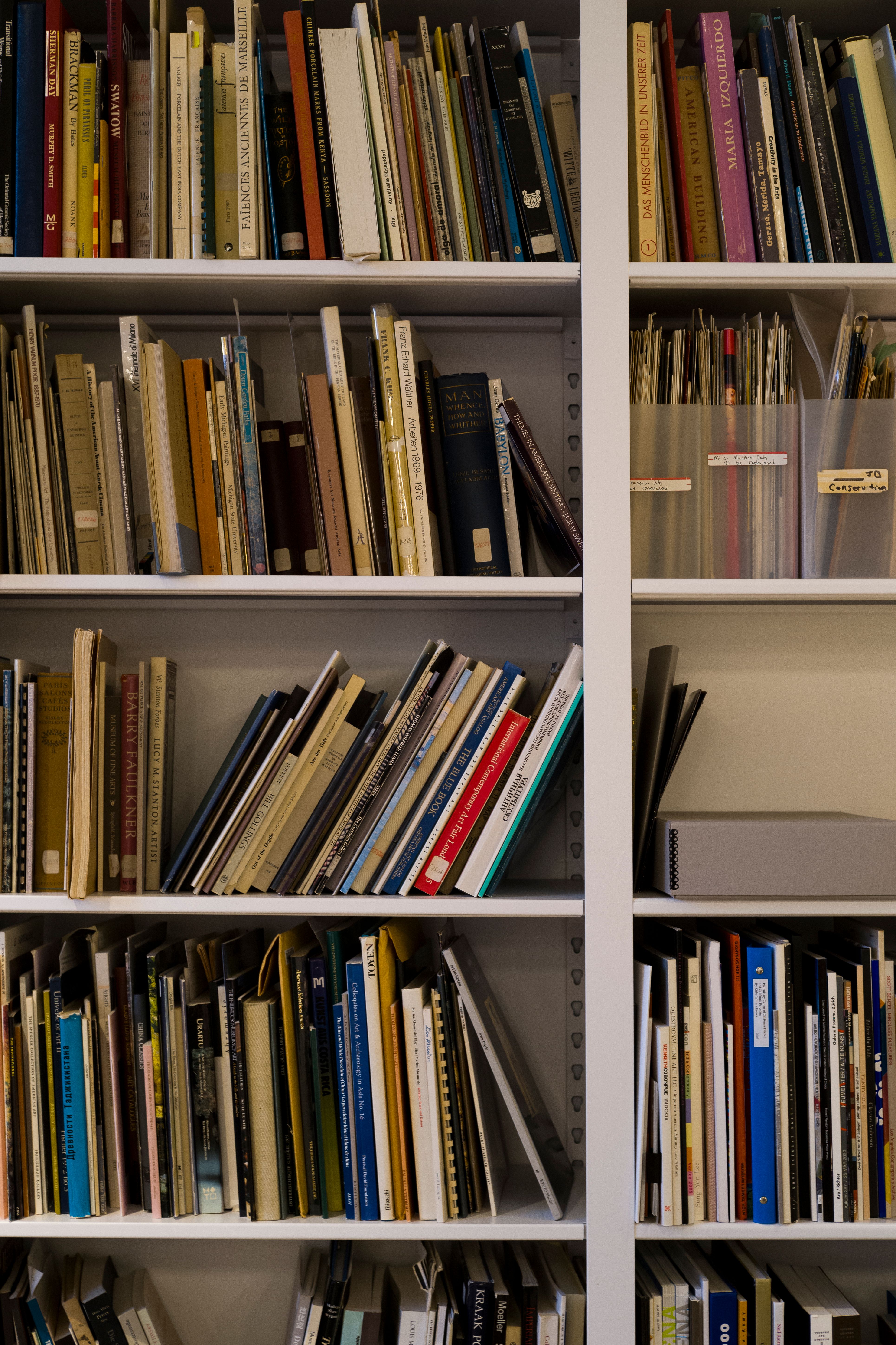A close-up of white bookshelves filled with art books, exhibition catalogues, and clear file boxes labeled by hand. Spines of books are arranged upright and at angles, creating a textured, archival feel.