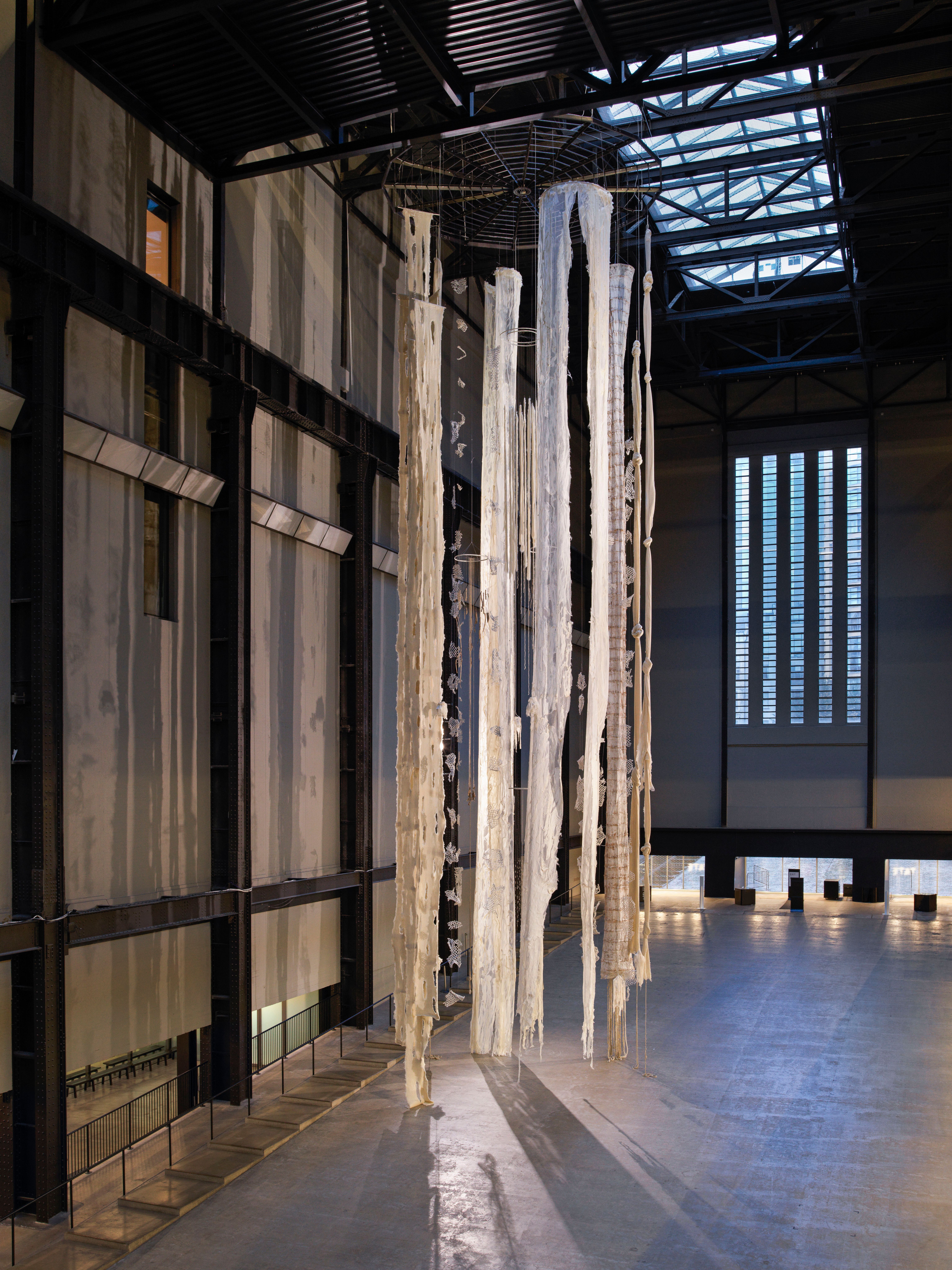 An enormous windchime installation fills a hall, adorned with long, tattered white materials.