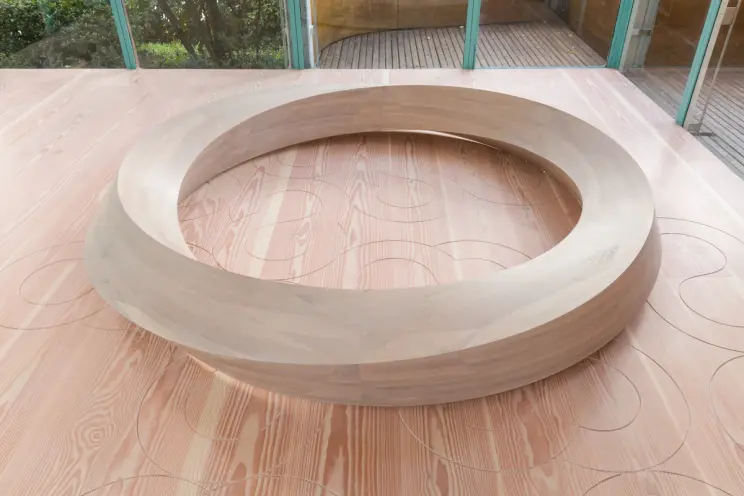 A close-up view of a circular, wooden bench on a light wooden floor with intricate patterns, situated next to large glass windows.