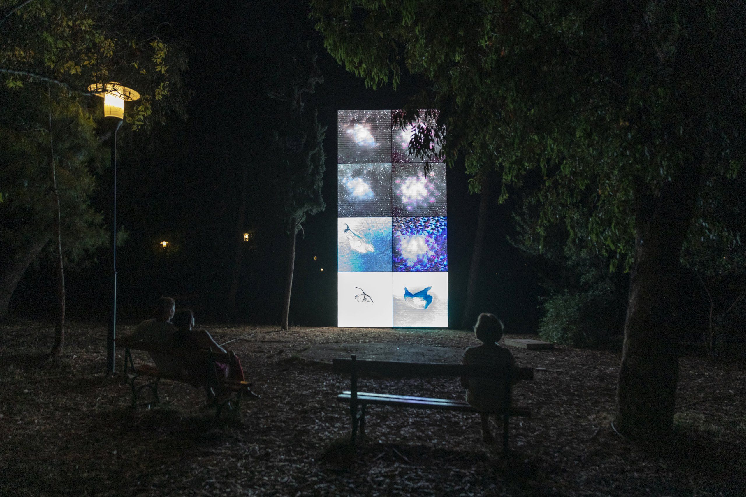 People sitting on benches at night in a park, watching a large digital screen displaying abstract cosmic and underwater scenes.