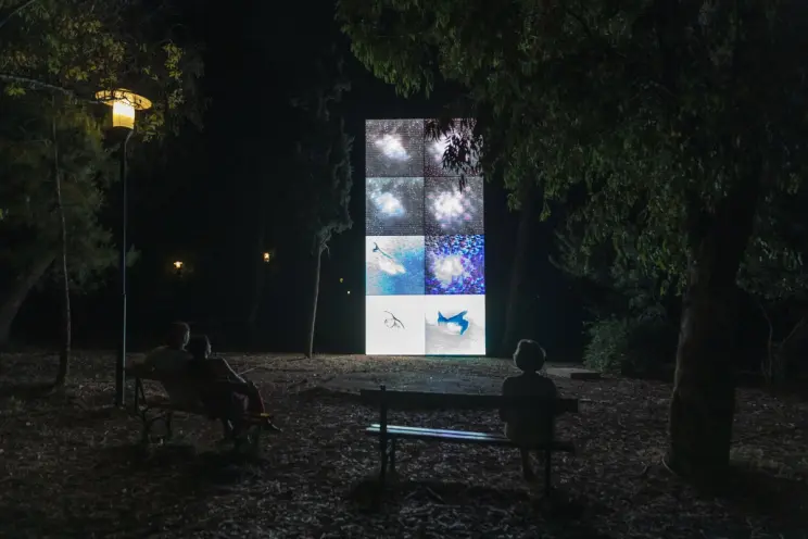People sitting on benches at night in a park, watching a large digital screen displaying abstract cosmic and underwater scenes.
