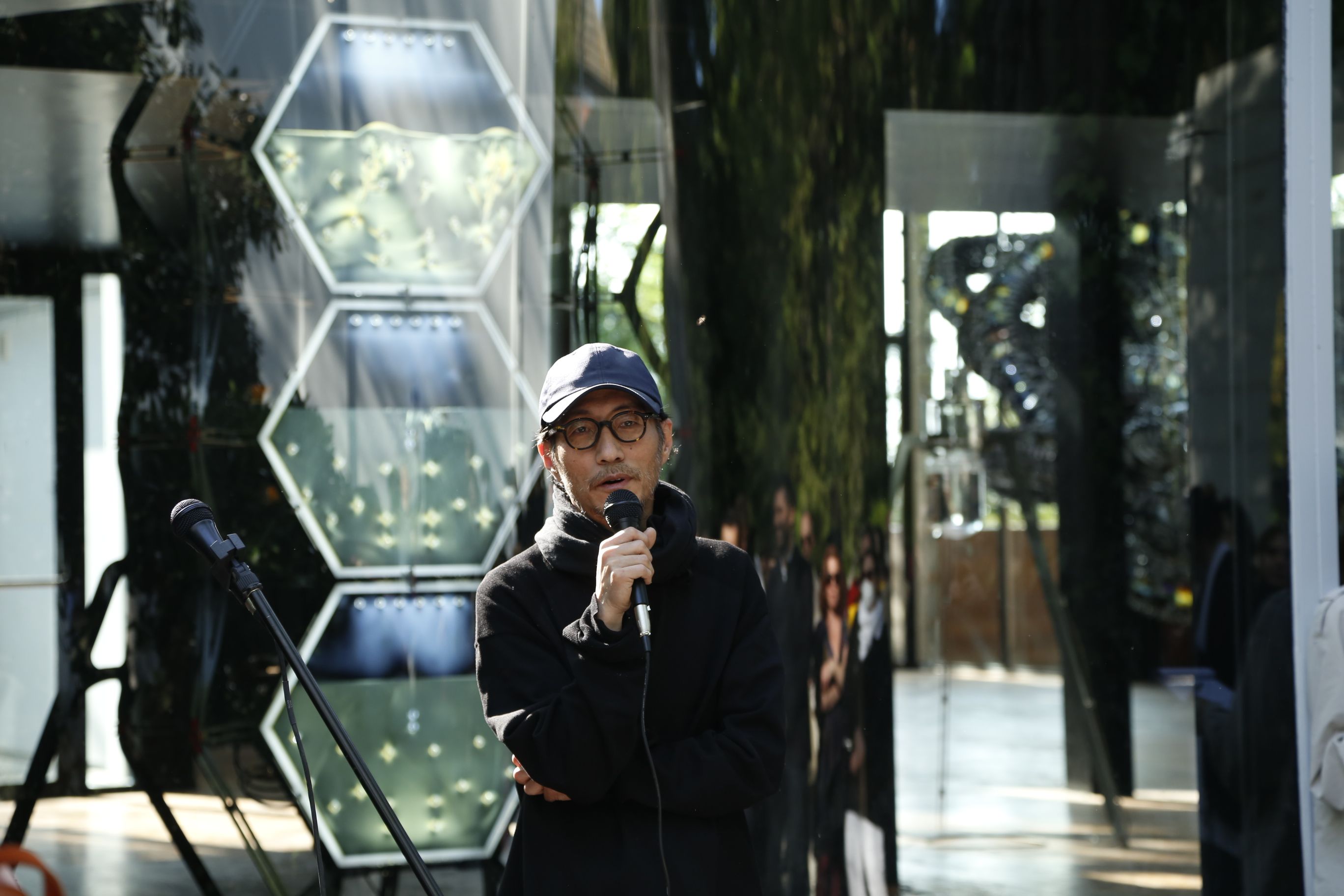 Yunchul Kim, holding a microphone and appearing to speak. Behind them, within glass encasements, various art pieces can be...