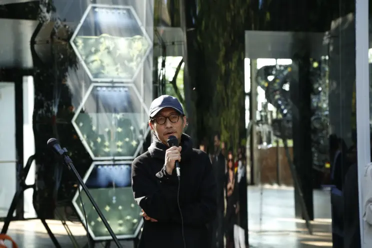 Yunchul Kim, holding a microphone and appearing to speak. Behind them, within glass encasements, various art pieces can be...