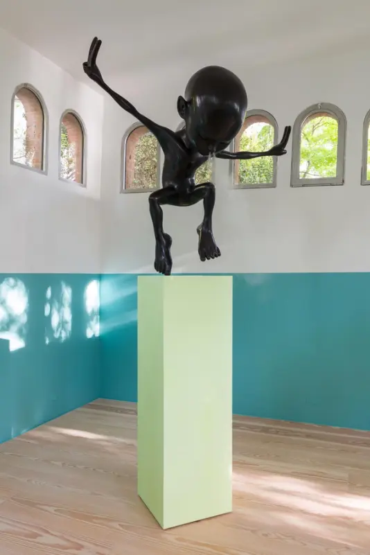 Vertical view of an art exhibit with a glossy black humanoid sculpture on a green rectangular column, against a teal and white dual-toned wall with arched windows and a light wooden floor.