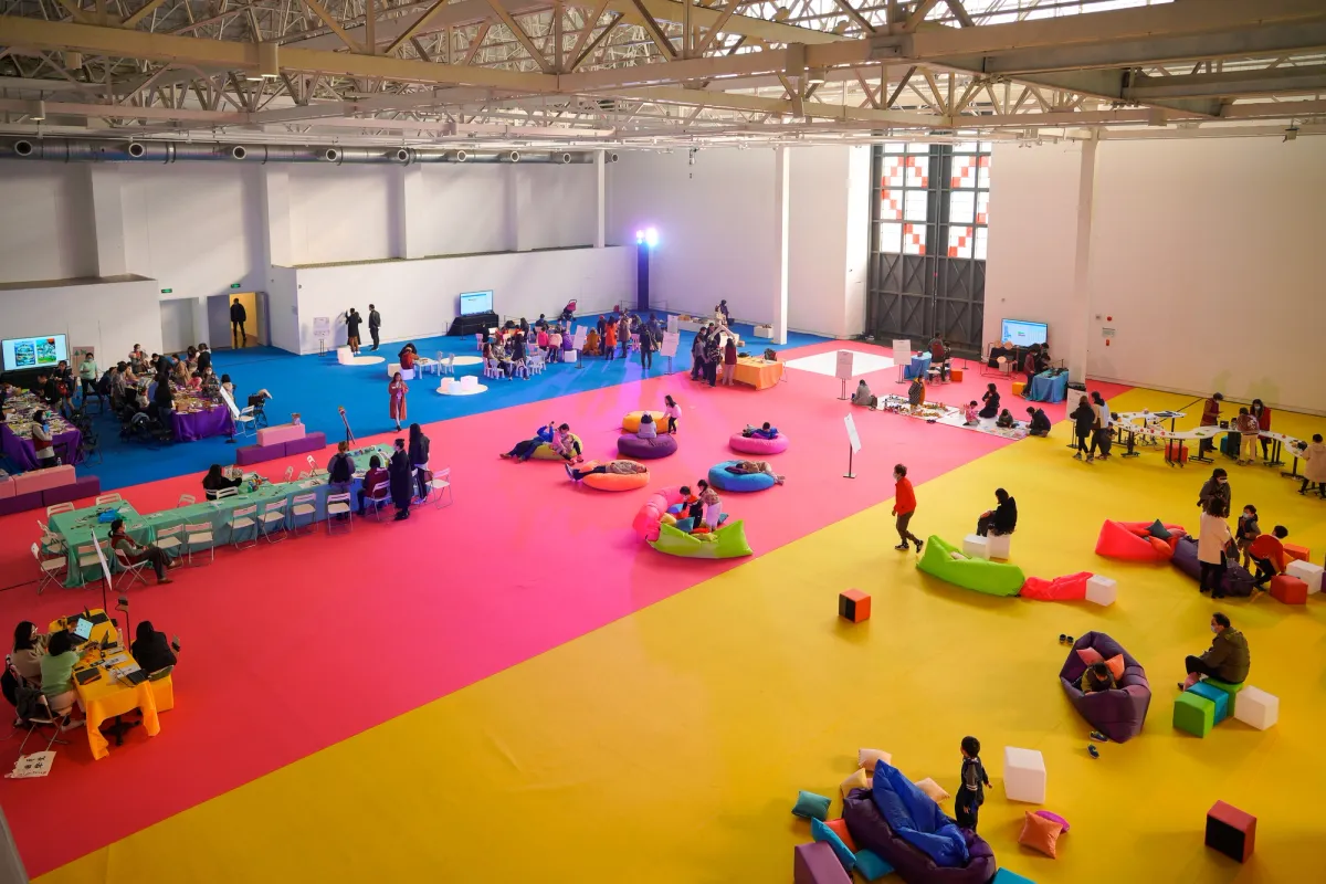 A spacious hall with a vibrant, multicolored floor hosting a family event. People in small groups are engaged in various activities, sitting at long tables with chairs or lounging on colorful bean bags. The viewpoint is a bird's eye perspective.