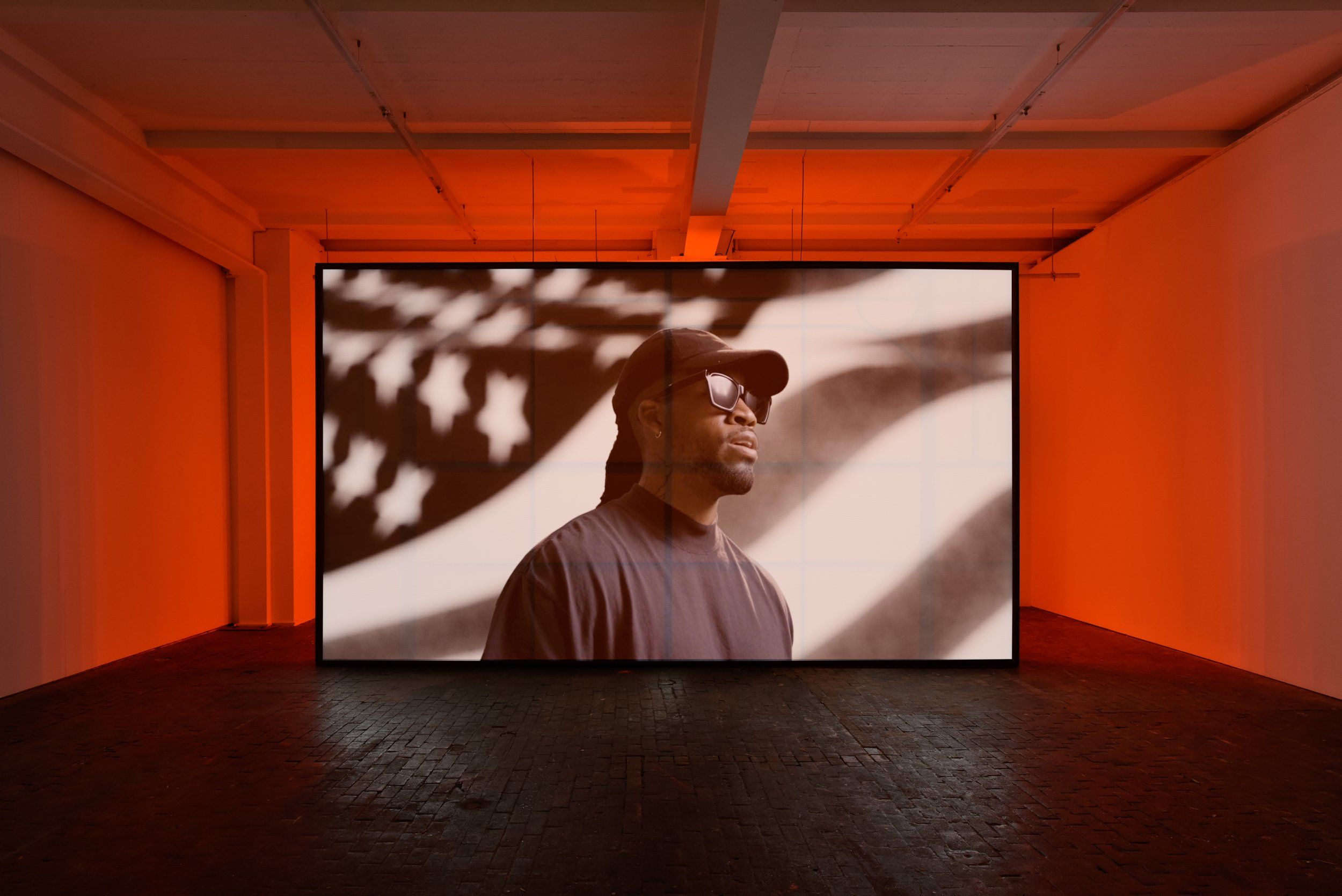 A person wearing sunglasses and a cap is projected on a large screen in a room illuminated by orange lighting, with an American flag as the background.